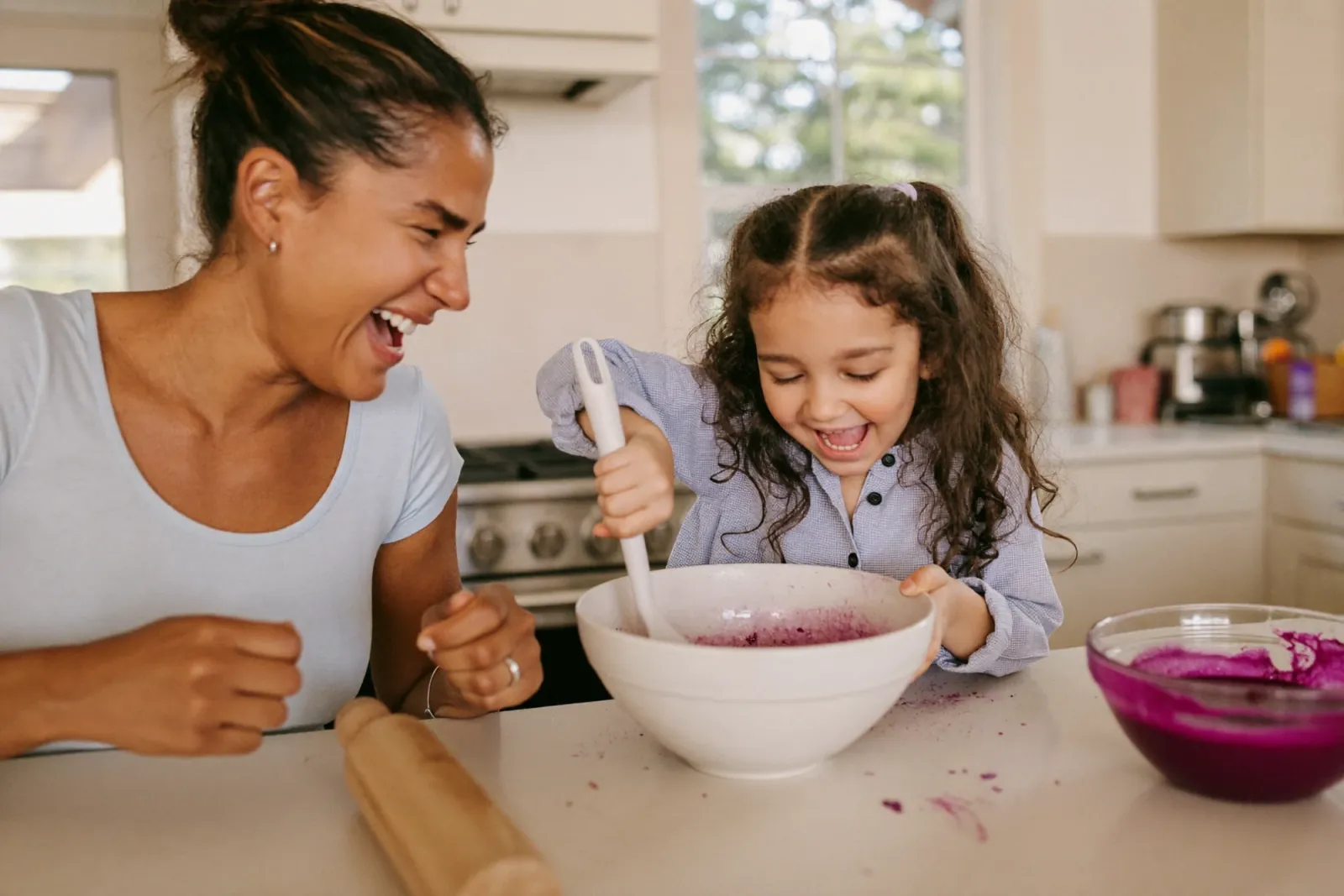 A mother and daughter laughing while mixing purple ube batter in a white bowl